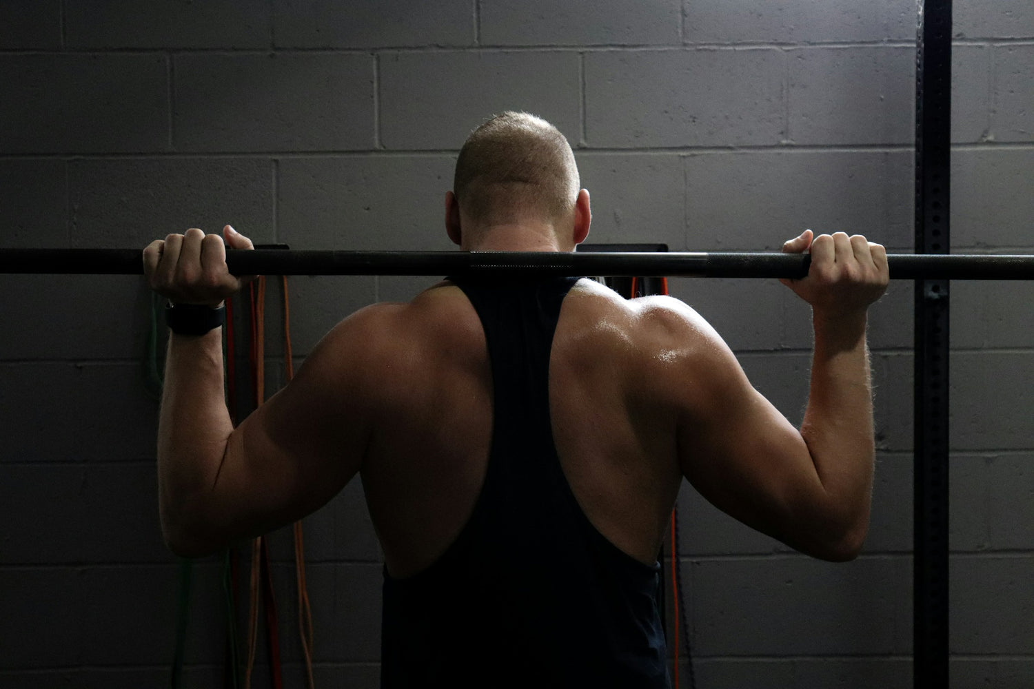 Close-up of a man's back doing pull-ups in a dark, moody gym with concrete walls